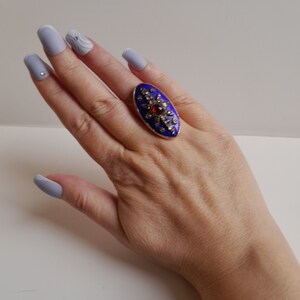 May include: A gold ring with a blue enamel oval setting. The oval is decorated with a starburst design of small diamonds and a single red gemstone in the center.