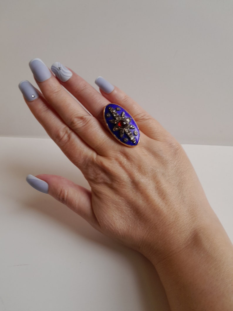 May include: A gold ring with a blue enamel oval setting. The oval is decorated with a starburst design of small diamonds and a single red gemstone in the center.