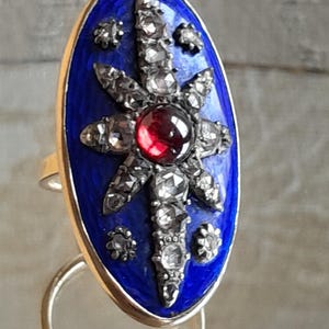 May include: A silver ring with a blue enamel oval setting. The setting features a red gemstone surrounded by smaller white gemstones in a starburst pattern.
