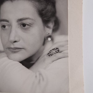 May include: Black and white portrait photograph of a woman with short, dark hair. She is wearing a ring with a large, ornate stone. The photo is signed "Lucie Dubail 1958."