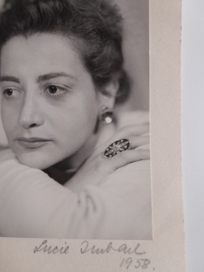 May include: Black and white portrait photograph of a woman with short, dark hair. She is wearing a ring with a large, ornate stone. The photo is signed "Lucie Dubail 1958."