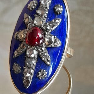 May include: A vintage ring with a blue enamel oval setting featuring a starburst design with a red gemstone in the center and surrounded by small diamonds. The ring is made of gold.
