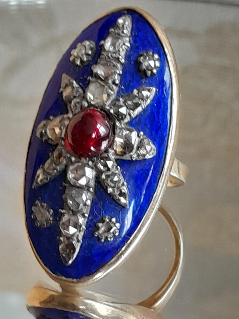 May include: A vintage ring with a blue enamel oval setting featuring a starburst design with a red gemstone in the center and surrounded by small diamonds. The ring is made of gold.