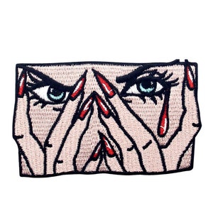 May include: Embroidered patch featuring a stylised image of a woman's face with red nail polish and red tears. Her eyes are wide open and her hands are covering her face.