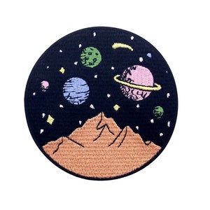 May include: A black and white embroidered patch with a mountain range in the foreground and a dark blue, pink, and green planets in the background. The planets are surrounded by white stars on a black background.