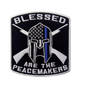Può includere: Toppa ricamata in bianco e nero con un elmo spartano, fucili incrociati e il testo "Blessed Are The Peacemakers". L'elmo ha una linea blu sul lato e la bandiera americana è visibile dietro.