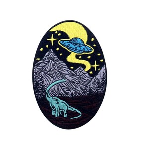 May include: A black and white embroidered patch with a green dinosaur and a blue UFO flying over a purple mountain range. The patch has a yellow star and a yellow moon in the background.