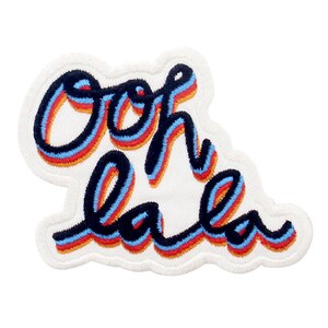 Könnte beinhalten: Ein weißer Stoff-Patch mit dem Schriftzug "Ooh la la", der in schwarzem, blauem, rotem und orangefarbenem Garn gestickt ist. Die Buchstaben sind schwarz umrandet und haben einen 3D-Effekt.