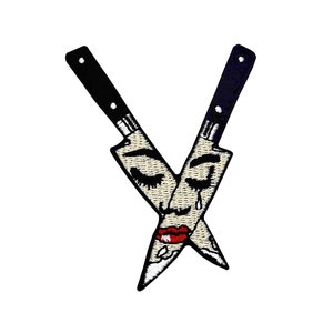 May include: Two black kitchen knives crossed over each other, forming a face with closed eyes, red lips, and a single teardrop. The knives are embroidered with a detailed design.