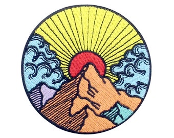 Puede incluir: Parche bordado con un paisaje montañoso y un sol naciente. El diseño incluye un sol rojo, rayos amarillos, nubes azules y una cordillera en tonos naranja, morado y marrón, todo dentro de un borde circular negro.