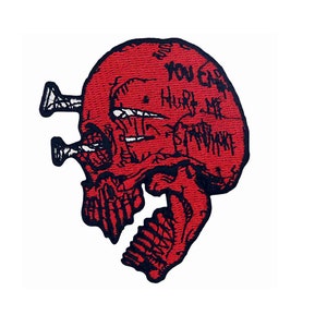 Puede incluir: Parche rojo bordado con forma de calavera con costuras negras y el texto "You Can't Hurt Me" escrito en la calavera. La calavera tiene dos clavos que sobresalen de la parte superior.