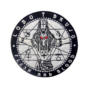 Pode incluir: Etiqueta bordada em preto e branco com um design circular. O centro apresenta uma figura branca com olhos vermelhos, cercada por formas geométricas. O círculo externo diz "LOBO Y DRUJO - FLESH AND BLOOD".