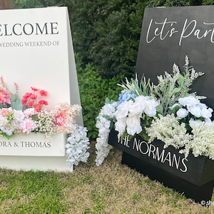 May include: Two white wooden A-frame signs with floral arrangements. The sign on the left says "Welcome to the wedding weekend of Alexandra & Thomas". The sign on the right says "Let's Party" and "The Normans".