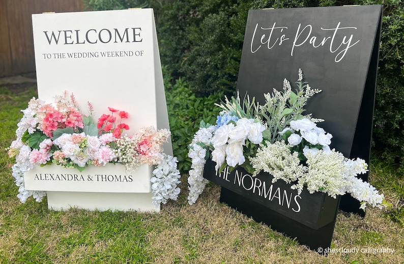 May include: Two white wooden A-frame signs with floral arrangements. The sign on the left says "Welcome to the wedding weekend of Alexandra & Thomas". The sign on the right says "Let's Party" and "The Normans".