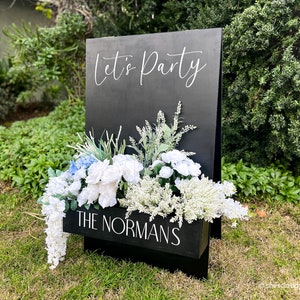 May include: Black wooden sign with the text "Let's Party" in white calligraphy. The sign has a planter box with white and blue flowers and the text "The Normans" in white.
