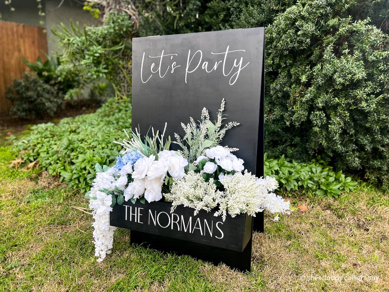 May include: Black wooden sign with the text "Let's Party" in white calligraphy. The sign has a planter box with white and blue flowers and the text "The Normans" in white.