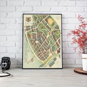 Op de afbeelding: Een ingelijste kaart van Riga, Letland, met een zwarte rand. De kaart toont een gedetailleerde illustratie van de indeling van de stad, inclusief straten, gebouwen en groene ruimtes. Het woord "RIGA" staat bovenaan gedrukt. Een vintage camera staat links.