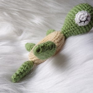 May include: A crocheted green and beige turtle toy lying on a white fluffy surface. The turtle has a white face with black eyes and a small smile.