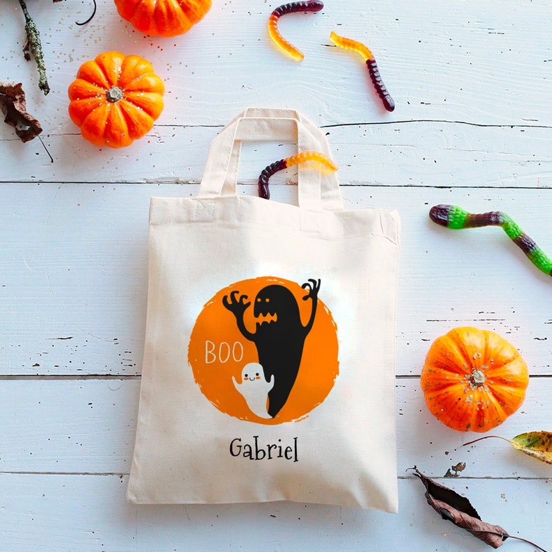 Op de afbeelding: Een witte canvas tote bag met een zwart-wit spookontwerp. De tas heeft een oranje cirkel met het woord "BOO" en de naam "Gabriel" erop gedrukt.