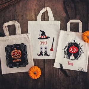 Op de afbeelding: Drie herbruikbare Halloween-tasjes met gepersonaliseerde namen. De tassen zijn wit met zwarte en oranje ontwerpen. De eerste tas heeft een heks met een zwarte hoed en de naam "Emma". De tweede tas heeft een jack-o-lantern met de naam "Charlie". De derde tas heeft een jack-o-lantern in een frame met de naam "Tom".