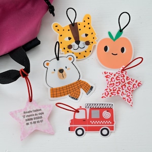 Peut inclure: Un ensemble de six étiquettes de sac décoratives présentant divers motifs, dont un visage de léopard jaune, une pêche orange, un ours blanc avec une écharpe à carreaux, un camion de pompiers rouge et deux étiquettes en forme d'étoile avec du texte.