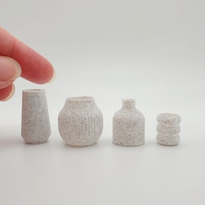 Puede incluir: Conjunto de cuatro jarrones de cerámica blanca con textura moteada. Los jarrones son de diferentes formas y tamaños, incluyendo un cilindro alto, un jarrón redondo, una botella corta y un cilindro pequeño.