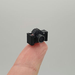 Puede incluir: Una cámara negra en miniatura con una lente plateada. La cámara está sentada en un dedo.