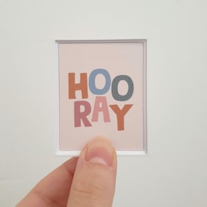 May include: A small framed print with the word "HOORAY" in colourful, block letters. The letters are in shades of brown, blue, pink, and yellow against a light pink background. The frame is white.