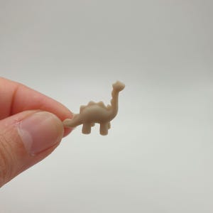 May include: A small, beige plastic dinosaur figurine. The dinosaur is standing on all fours with its tail curled up behind it. It has a long neck and a small head.