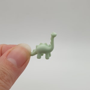 May include: A small, light green plastic dinosaur figurine. The dinosaur is standing on its four legs with its tail curled up behind it.