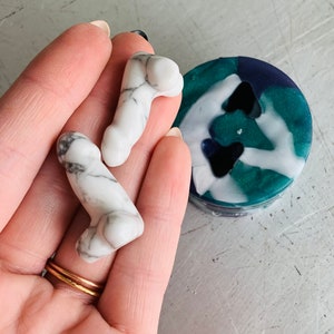 Puede incluir: Dos esculturas fálicas de mármol blanco y gris se sostienen en una mano, con un pequeño objeto redondo, turquesa, blanco y azul oscuro en el fondo. Las esculturas tienen una forma detallada y realista.