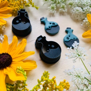 Puede incluir: Cuatro cortadores de galletas de botella de poción negra y verde sobre una superficie de madera con flores amarillas y blancas.