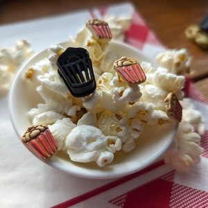 Peut inclure: Un bol blanc rempli de popcorn et décoré de petits boutons en forme de popcorn. Les boutons sont rouges, blancs et noirs.