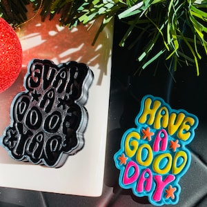 Puede incluir: Dos cortadores de galletas, uno negro y otro azul. El cortador negro tiene la forma de las palabras "Have a good day" en una fuente retro. El cortador azul tiene la forma de las palabras "Have a good day" en una fuente retro con acentos amarillos, rosados y naranjas.