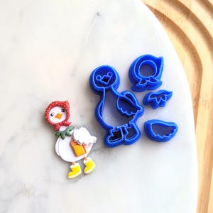 May include: Blue plastic cookie cutters shaped like a hen with a red bonnet, a chick, and a wing. The cutters are on a white marble surface.