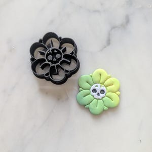 May include: A black plastic cookie cutter shaped like a flower with a skull in the center. Next to it is a green and yellow fondant cookie with a white skull in the center.