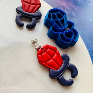 May include: Red and navy blue diamond ring earrings and a blue ring-shaped cutter. The earrings feature a red, faceted diamond shape atop a navy blue ring setting. The cutter is a blue plastic mold of the ring.