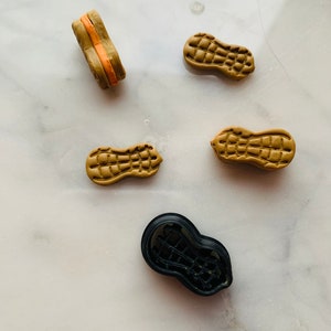 以下が含まれることがあります： ピーナッツの形をしたクッキー型が5つあり、4つは茶色で1つは黒です。茶色の型は、ピーナッツの殻のような質感のある表面をしています。黒い型は滑らかで、茶色の型と似た形をしています。