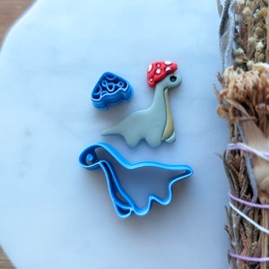 May include: A set of three blue plastic dinosaur cookie cutters. One cutter is a standing dinosaur with a red mushroom on its head. The other two cutters are of a lying dinosaur.