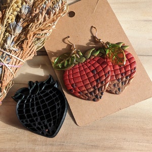 May include: A pair of red and gold strawberry earrings with green leaves. The earrings are on a brown card with a black strawberry cutout.