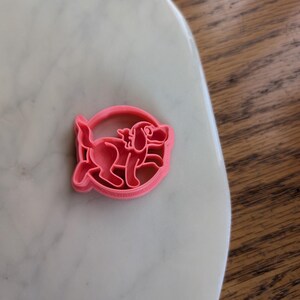 May include: A pink plastic cookie cutter in the shape of a cartoon dog inside a circle. The dog is depicted with floppy ears, a tail, and a smiling expression. The cutter is on a white marble surface.