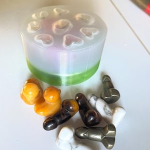Puede incluir: Una colección de pequeñas esculturas con forma fálica en varios colores y materiales, incluyendo ámbar, negro, blanco y acabados metálicos. Un molde cilíndrico con cavidades en forma de corazón.