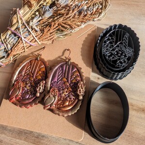 May include: A pair of gold and purple owl earrings with a forest scene. The earrings are on a brown background with a black oval and a black circular cookie cutter.
