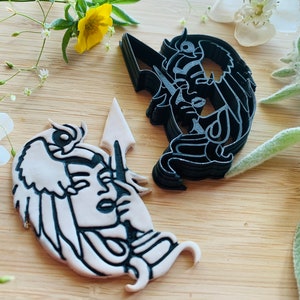 May include: Two black and white cookie cutters in the shape of a woman's face. The cookie cutters are on a wooden surface.