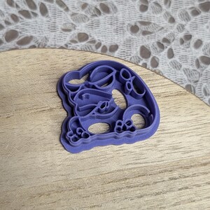 May include: A purple plastic cookie cutter shaped like a cartoon character. The cutter has a detailed design with rounded features and a scalloped edge. The cutter is on a wooden surface.