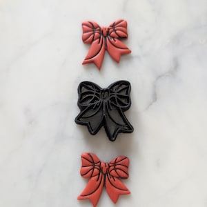 Puede incluir: Tres elementos en forma de lazo. Dos son de color coral y uno es un cortador de galletas negro. Los lazos tienen líneas detalladas y están dispuestos verticalmente sobre una superficie de mármol blanco. El cortador de galletas está en el medio.