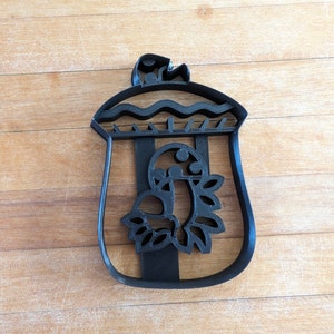 May include: Black plastic cookie cutter in the shape of a bag with a bird and floral design. The bag has vertical stripes and a wavy top edge. The bird is in the center of the bag, surrounded by a floral wreath.