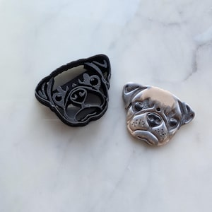 Peut inclure: Un emporte-pièce noir en forme de carlin à côté d'un pendentif argenté en forme de carlin. L'emporte-pièce est en plastique et présente des détails complexes. Le pendentif est en métal et représente un visage de carlin.