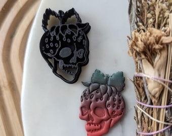 May include: Two black and red 3D printed cookie cutters shaped like strawberry skulls. The cutters are on a white surface.