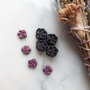 May include: Five black metal rose-shaped buttons with a raised design. Four of the buttons are arranged in a cross shape, with the fifth button placed on top of the cross. There are five smaller purple rose-shaped buttons scattered around the larger buttons.